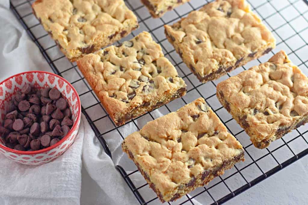 Six chocolate chip cookie bars, made using a cake mix recipe, cool on a wire rack beside a small bowl of chocolate chips on a white surface.