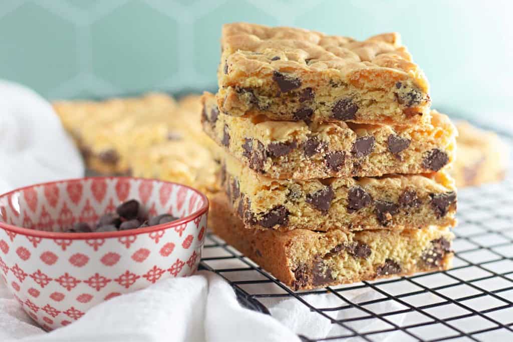 A stack of chocolate chip cookie bars made from cake mix recipes sits on a cooling rack next to a red and white patterned bowl filled with chocolate chips.