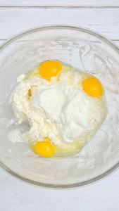 A glass bowl containing three raw eggs, cream cheese, and sour cream on a white wooden surface.