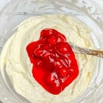 A glass bowl containing creamy white mixture with a bright red fruit topping and a spoon, placed on a white surface.