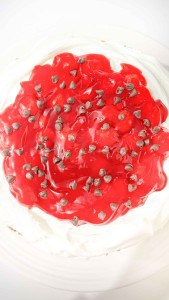 A cake topped with white whipped cream, red fruit glaze, and sprinkled with chocolate chips.