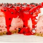 A cherry-topped cheesecake with a graham cracker crust, pink filling, whipped cream, chocolate chips, and a slice removed, displayed on a white plate.