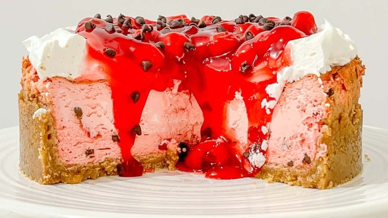 A cherry-topped cheesecake with a graham cracker crust, pink filling, whipped cream, chocolate chips, and a slice removed, displayed on a white plate.