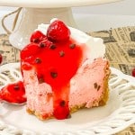 A slice of cherry cheesecake with whipped cream, cherry topping, and chocolate chips on a white plate, surrounded by fresh cherries.