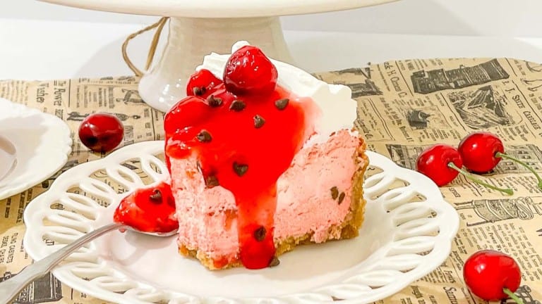 A slice of cherry cheesecake with whipped cream, cherry topping, and chocolate chips on a white plate, surrounded by fresh cherries.