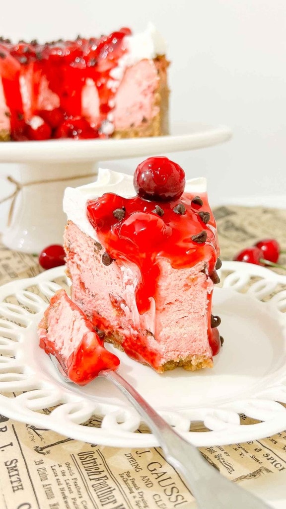 A slice of cherry cheesecake with whipped cream, cherry topping, and chocolate chips sits on a white plate. A fork cuts into the slice. The background shows the rest of the cake.