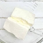 Two blocks of cream cheese in a clear glass mixing bowl on a white wooden surface.