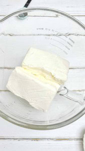 Two blocks of cream cheese in a clear glass mixing bowl on a white wooden surface.