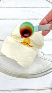 A hand pours vanilla extract from a measuring spoon into a glass bowl containing two blocks of cream cheese.