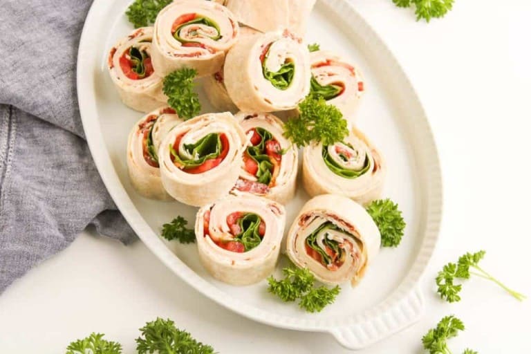 A white plate with tortilla pinwheels filled with deli meat, cheese, lettuce, and tomatoes, garnished with fresh parsley.