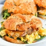 A croissant sandwich filled with scrambled eggs, spinach, cheese, and sausage is served on a white plate.