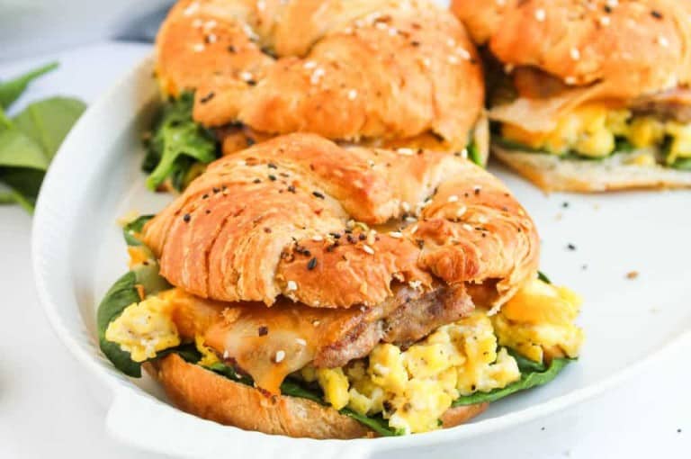 A croissant sandwich filled with scrambled eggs, spinach, cheese, and sausage is served on a white plate.