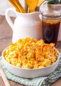A white bowl filled with creamy potato salad topped with paprika sits on a green patterned napkin beside a glass of iced drink and wooden utensils.