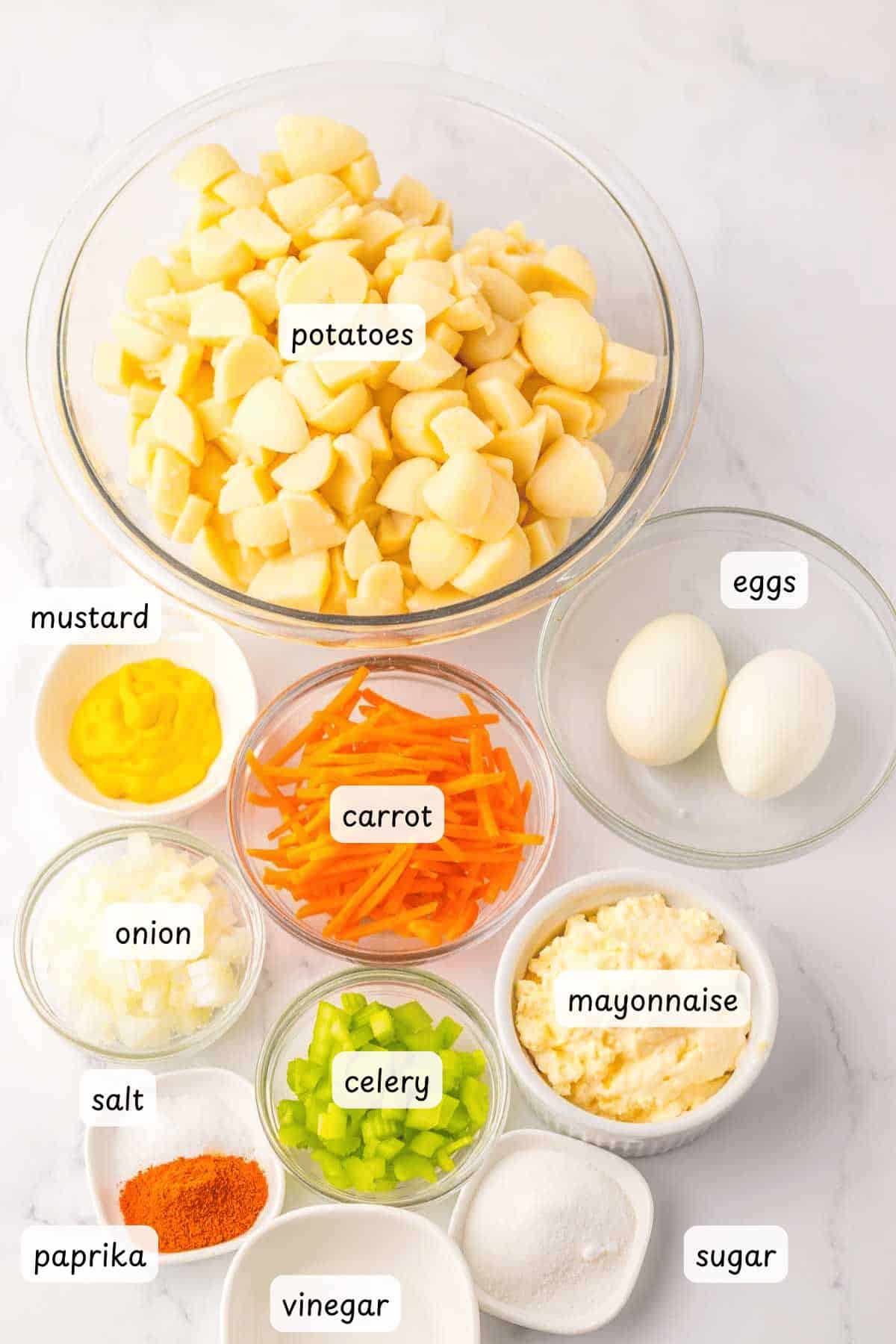 Various ingredients for potato salad are displayed in bowls on a white surface, including potatoes, eggs, carrot, onion, celery, mayonnaise, mustard, salt, paprika, vinegar, and sugar.