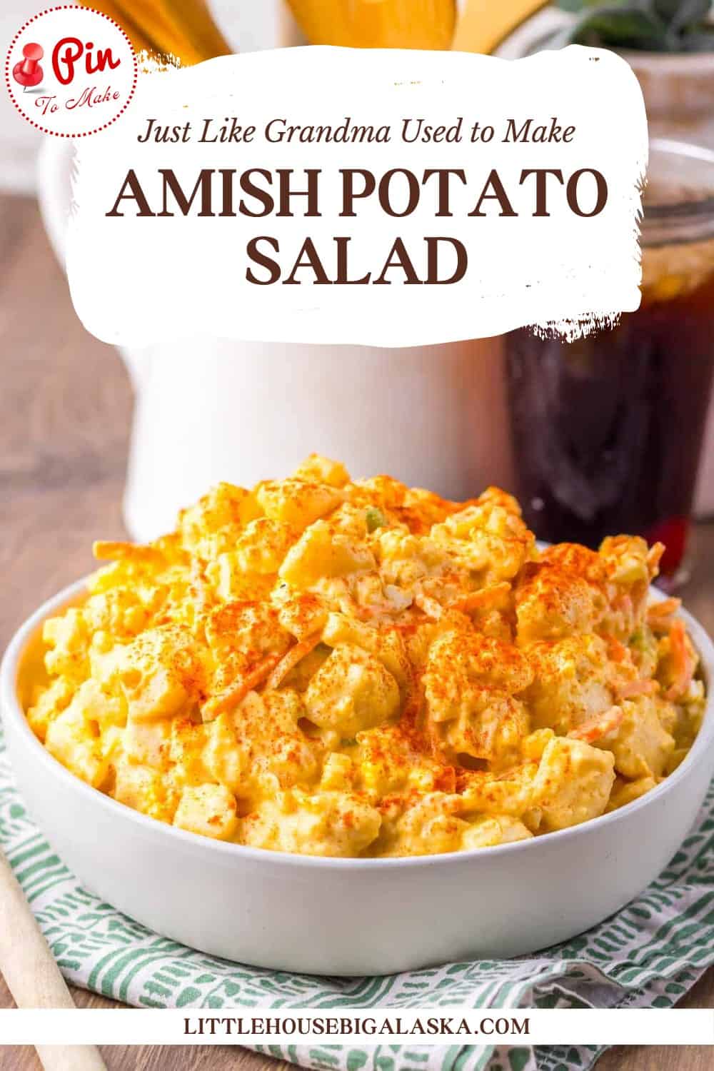 A bowl of Amish potato salad topped with paprika sits on a striped cloth, with a website link below and a glass of iced tea in the background.
