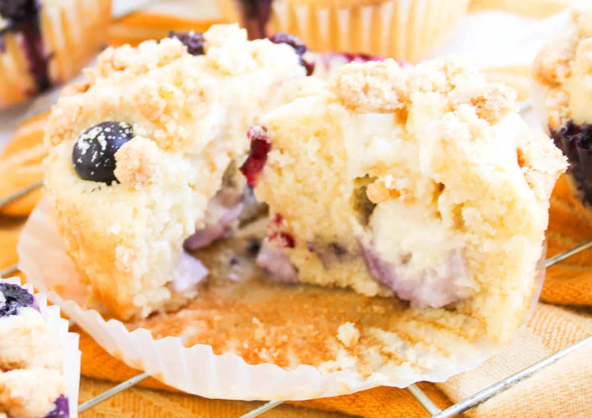 A close-up of Blueberry Cheesecake Muffins cut in half, showing a creamy filling and blueberries inside, with a crumbly topping, resting on a muffin liner.