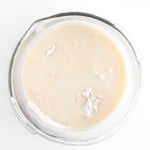 A glass mixing bowl containing a pale, creamy batter for Blueberry Cheesecake Muffins, with patches of unmixed flour on the surface, set against a white background.