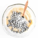 A glass mixing bowl with batter and flour-coated blueberries being stirred for Blueberry Cheesecake Muffins using a wooden spatula on a white background.