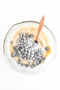 A glass mixing bowl with batter and flour-coated blueberries being stirred for Blueberry Cheesecake Muffins using a wooden spatula on a white background.