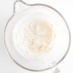 A glass mixing bowl containing whipped cream cheese on a white surface, perfect for making Blueberry Cheesecake Muffins.