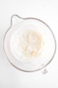 A glass mixing bowl containing whipped cream cheese on a white surface, perfect for making Blueberry Cheesecake Muffins.
