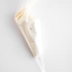 A clear plastic piping bag filled with white icing or cream, perfect for topping fresh blueberry cheesecake muffins, placed on a white surface.