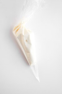 A clear plastic piping bag filled with white icing or cream, perfect for topping fresh blueberry cheesecake muffins, placed on a white surface.