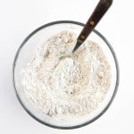 A glass bowl contains a mixture of flour and other dry ingredients for Blueberry Cheesecake Muffins, being stirred with a metal fork with a dark wooden handle.