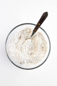 A glass bowl contains a mixture of flour and other dry ingredients for Blueberry Cheesecake Muffins, being stirred with a metal fork with a dark wooden handle.