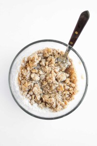 A glass bowl filled with a crumbly brown sugar and flour mixture, perfect for topping Blueberry Cheesecake Muffins, with a fork resting inside, on a white background.