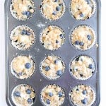 A muffin tray with twelve paper liners filled with unbaked Blueberry Cheesecake Muffins batter and a generous streusel topping.