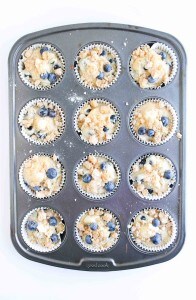 A muffin tray with twelve paper liners filled with unbaked Blueberry Cheesecake Muffins batter and a generous streusel topping.