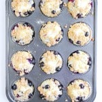 A muffin tin with twelve Blueberry Cheesecake Muffins, each topped with crumbly streusel and some showing juicy blueberries peeking through.