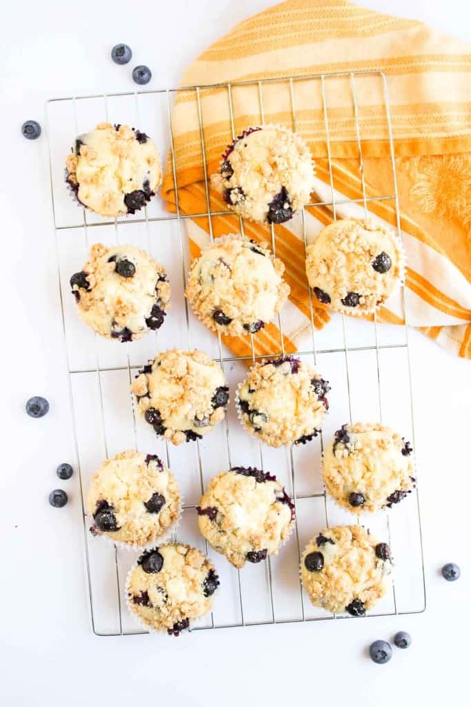 Twelve Blueberry Cheesecake Muffins with crumb topping cool on a wire rack, surrounded by a few loose blueberries and a yellow-striped kitchen towel.