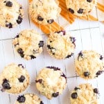 Nine Blueberry Cheesecake Muffins with crumb topping are cooling on a wire rack, with a yellow-striped kitchen towel and scattered blueberries nearby.