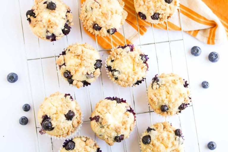 Nine Blueberry Cheesecake Muffins with crumb topping are cooling on a wire rack, with a yellow-striped kitchen towel and scattered blueberries nearby.
