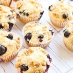 Blueberry Cheesecake Muffins with crumb topping in paper liners are cooling on a wire rack, with a yellow striped cloth and loose blueberries in the background.