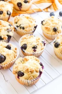 Blueberry Cheesecake Muffins with crumb topping in paper liners are cooling on a wire rack, with a yellow striped cloth and loose blueberries in the background.