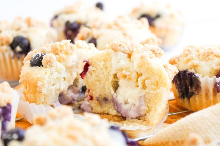 A close-up of Blueberry Cheesecake Muffins with crumb topping, one split open to reveal a creamy cheesecake filling and whole blueberries inside.