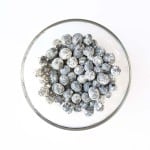 A glass bowl filled with blueberries coated in a layer of white flour, photographed from above on a white background&mdash;perfectly prepped for Blueberry Cheesecake Muffins.