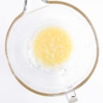 A glass mixing bowl containing melted butter and sugar, the perfect start for baking delicious Blueberry Cheesecake Muffins, sits on a white background.