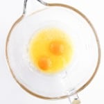 A clear glass mixing bowl containing two raw eggs and melted butter on a white background, perfect for starting your Blueberry Cheesecake Muffins recipe.