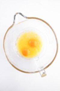 A clear glass mixing bowl containing two raw eggs and melted butter on a white background, perfect for starting your Blueberry Cheesecake Muffins recipe.
