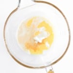 A glass mixing bowl containing milk, melted butter, yogurt, spices, and ingredients for Blueberry Cheesecake Muffins sits on a white surface.