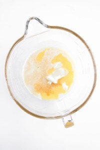 A glass mixing bowl containing milk, melted butter, yogurt, spices, and ingredients for Blueberry Cheesecake Muffins sits on a white surface.