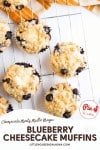 Six Blueberry Cheesecake Muffins with crumb topping are cooling on a wire rack, viewed from above. Text on image reads "Blueberry Cheesecake Muffins," showcasing these delicious homemade treats.