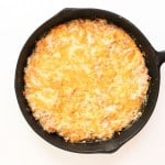 A cast iron skillet filled with a baked, golden-brown cheesy casserole on a white background.