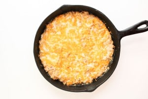 A cast iron skillet filled with a baked, golden-brown cheesy casserole on a white background.