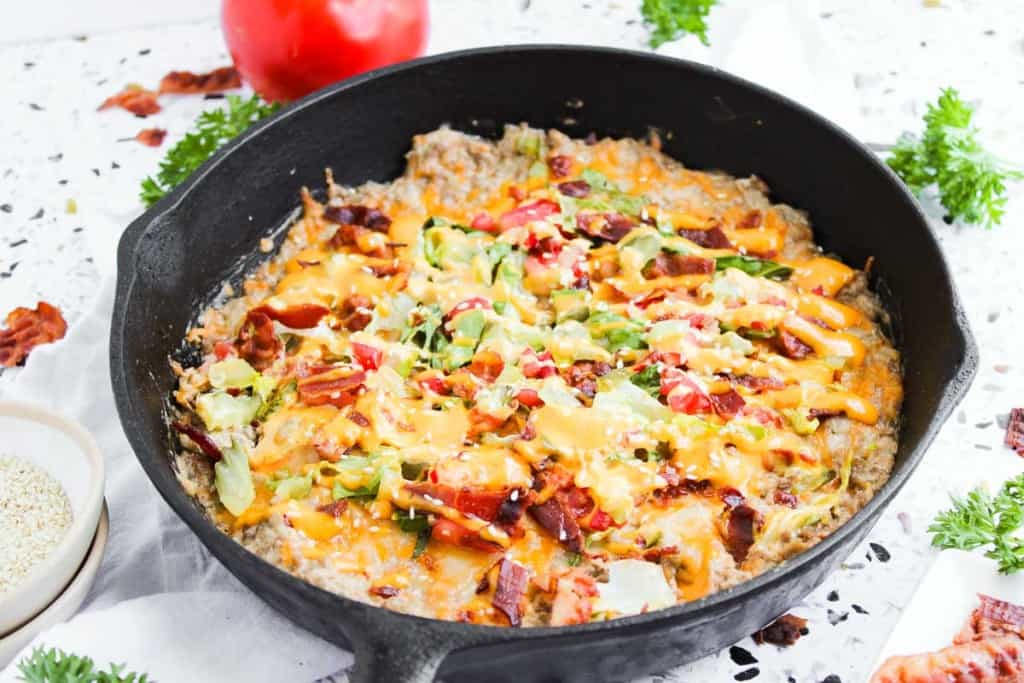 A cast iron skillet filled with a baked dish topped with melted cheese, chopped lettuce, tomatoes, and bacon pieces, surrounded by herbs and a tomato.