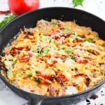 A cast iron skillet filled with a baked dish topped with melted cheese, chopped lettuce, tomatoes, and bacon pieces, surrounded by herbs and a tomato.